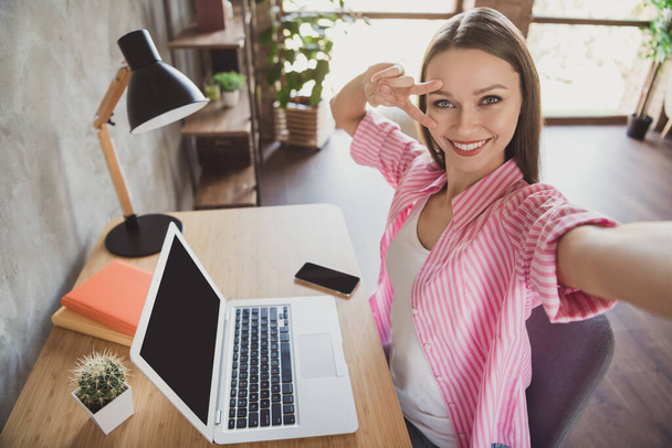 Фото позитивной каштановые волосы женщина сделать селфи камеры показывают V-знак сидеть стол домашний офис ноутбук в помещении дома - Фото, изображение
