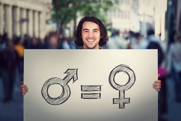 Gendergelijkheidsconcept als jongeman met een groot spandoek met mannelijk en vrouwelijk symbool, als demonstratieboodschap op een drukke straat. Seksrechten en discriminatie als belangrijke sociale kwestie - Foto, afbeelding