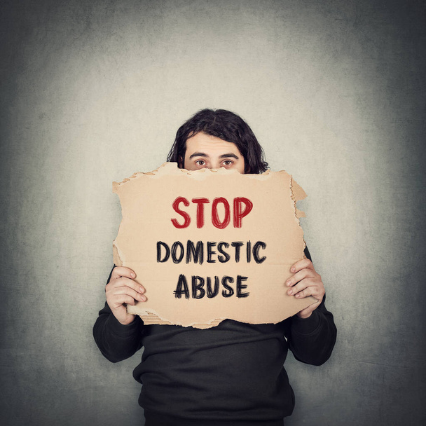 Stop huiselijk geweld. Bang man, slachtoffer van huiselijk geweld, met een kartonnen spandoek met sms-bericht geïsoleerd op grijze muur achtergrond. Huiselijk geweld als sociaal probleem - Foto, afbeelding