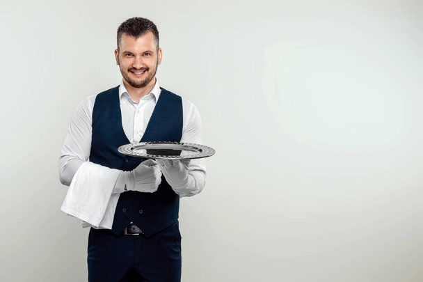 Handsome male waiter, in white shirt, holding a silver tray. The concept of serving staff serving customers in a restaurant - Фото, изображение