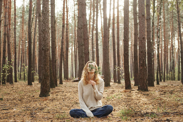  Caucasian woman faceless outdoors sitting on the ground. she covered her face with a spruce branch. The concept of unity with nature and well-being. - Photo, Image