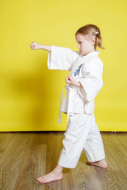 Portrait Of 5 year old caucasian Girl in kimono Practicing Karate Against yellow Background at home - Фото, изображение