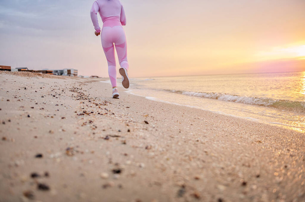 Частковий вид ззаду невпізнаваної жінки-бігунки в рожевому спортивному вбранні, що виконує біг на пляжі рано вранці
 - Фото, зображення