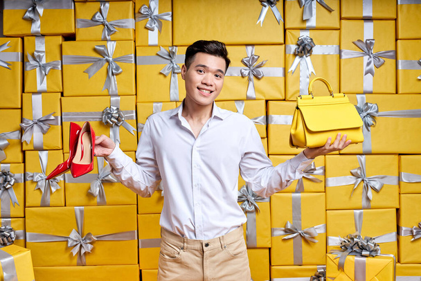 Asian man offers purchase options, holding shoes and handbag in hands - Fotografie, Obrázek