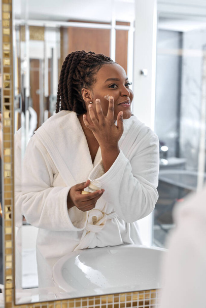 Pretty young African woman wearing white robe applying moisturizer on face looking at mirror - Foto, Imagem