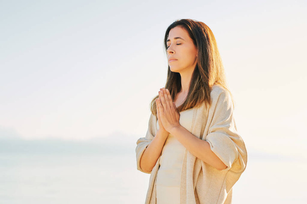 Beautiful spiritual woman meditating by the lake, wearing beige clothes - Photo, Image