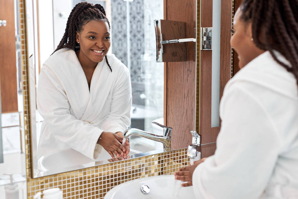 Black woman washing hands in modern bright bathroom, looking at mirror reflection - 写真・画像