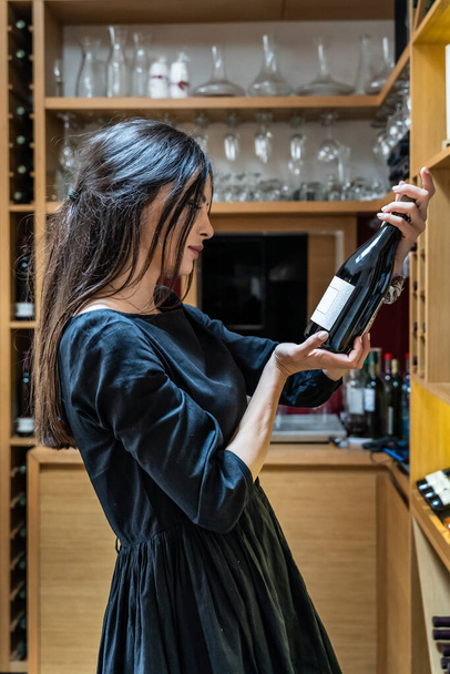 Retrato vista lateral de mujer caucásica adulta en vestido negro recogiendo la botella de vino en casa en la cocina de pie solo - Foto, Imagen