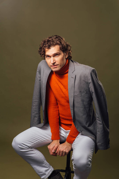 Full lengh portrait of a tall handsome man dressed in orange turtleneck, grey jacket and white jeans posing on the green background - Φωτογραφία, εικόνα