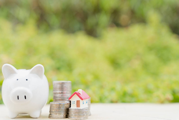 Piggy bank met huis op stapel munten op wazig groene natuurlijke achtergrond. Besparen van geld en investeringen. - Foto, afbeelding