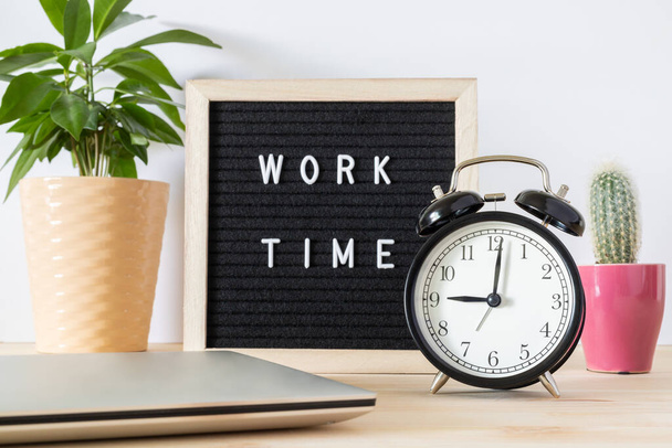 Classic vintage black alarm clock, laptop, potted plants and board sign 'work time' on wooden desk against white wall background. Time and work concept. Start of new working day. - Photo, Image
