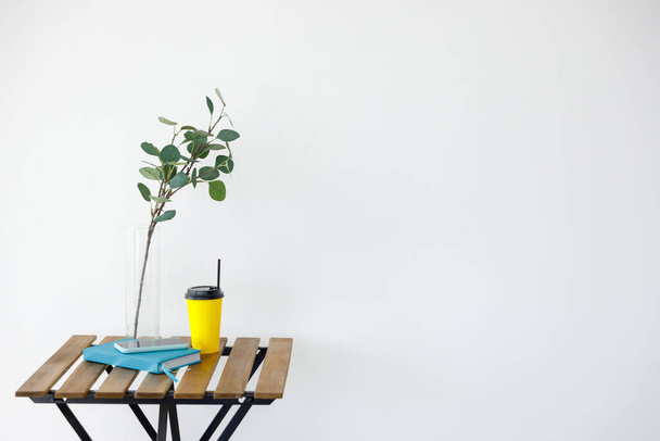 Wooden table against background of white wall. Vase with eucalyptus branch, yellow paper coffee cup and smartphone with notepad. Freelancer workplace - Fotografie, Obrázek