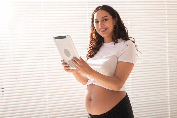 Счастливая беременная молодая красивая женщина разговаривает со своим мужем с помощью видео связи и планшета. Концепция общения и позитивное отношение во время беременности - Фото, изображение