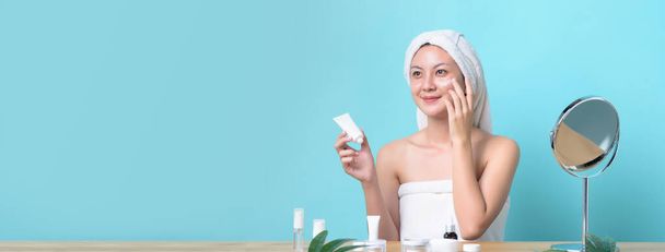 Beautiful Asian young woman smiling while applying white cream on her face with many packaging of facial cream and cosmetics on wooden table. Banner size with copy space light blue background. - Photo, Image