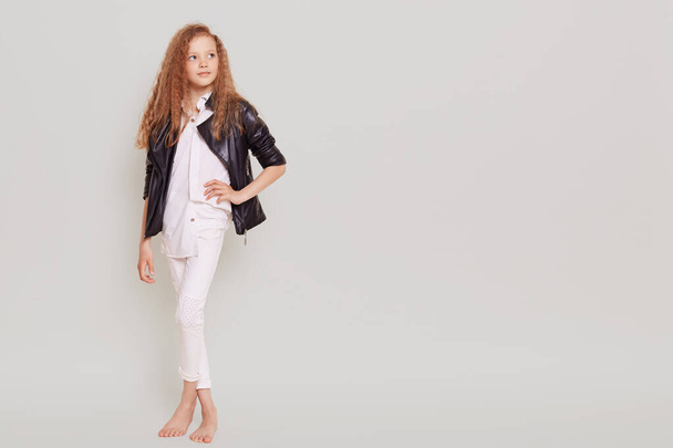 Full length portrait of confident little schoolgirl wearing white clothing and leather jacket looking away and keeping hand on hip, copy space, isolated over gray background. - Foto, afbeelding