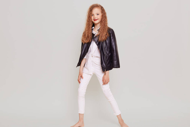 Full length portrait of confident schoolgirl wearing stylish attire and leather jacket, looking at camera, standing in trendy position, has blonde wavy hair, isolated over gray background. - 写真・画像