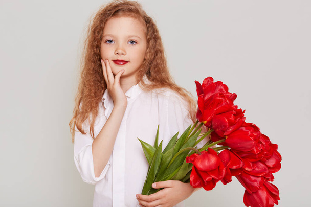 Charming pretty child girl wearing white clothing gets big bouquet of red tulips as present, looking at camera wit confident expression ,isolated over gray background. - 写真・画像