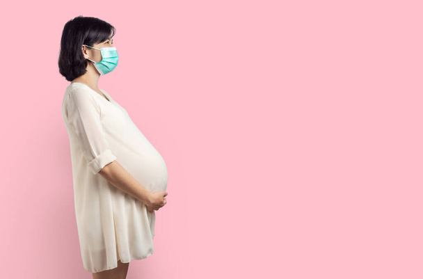 Feliz Sorrindo Asiático Mulher grávida vestindo máscara protetora em pé sobre fundo isolado rosa. Mulher expectante segurando sua barriga com espaço de cópia. - Foto, Imagem