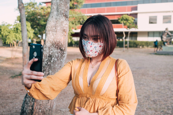 Een Aziatische dame met een gezichtsmasker neemt een selfie van zichzelf op een grasveld in de buurt van haar werkplek. - Foto, afbeelding