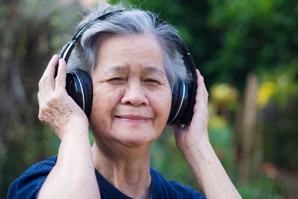 Oudere vrouw staat met een draadloze hoofdtelefoon te luisteren naar een favoriet lied, en glimlach gelukkig in de tuin. Aziatische senior vrouw, kort wit haar genieten van de muziek. Goed concept van geestelijke gezondheid - Foto, afbeelding