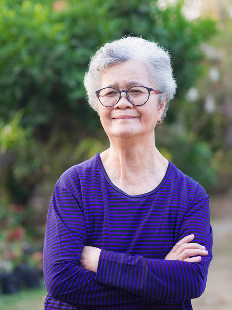 A portrait of an senior woman arms crossed and smiling while standing in a garden. Space for text. Concept of old people and healthcare - Photo, Image