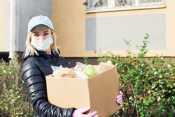 Eine freiwillige Helferin mit Schutzmaske geht mit einer Kiste voller Lebensmittel, Spenden, auf die Straße.. - Foto, Bild