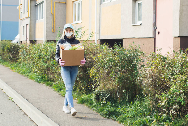εθελόντρια που φοράει προστατευτική μάσκα περπατάει στο δρόμο με ένα κουτί με ψώνια, δωρεές. - Φωτογραφία, εικόνα