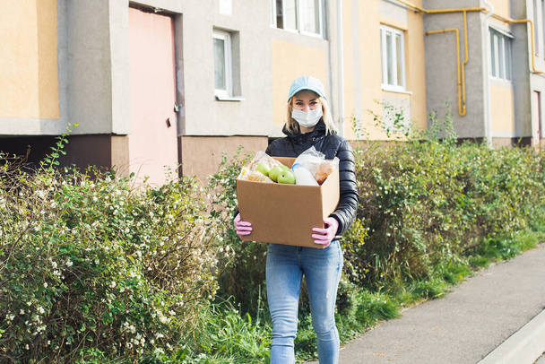 防護マスクを着た女性ボランティアが食料品箱を持って通りを歩き寄付をしています. - 写真・画像