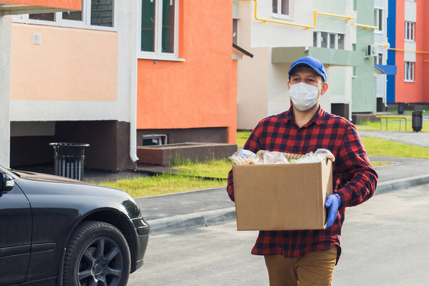 un voluntario masculino con una máscara protectora camina por la calle con una caja de comestibles, caridad. - Foto, imagen