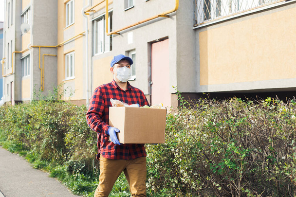 Ένας άντρας εθελοντής με προστατευτική μάσκα περπατάει στο δρόμο με ένα κουτί με ψώνια, φιλανθρωπία.. - Φωτογραφία, εικόνα