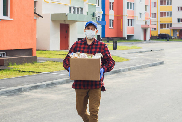 Ένας άντρας εθελοντής με προστατευτική μάσκα περπατάει στο δρόμο με ένα κουτί με ψώνια, φιλανθρωπία.. - Φωτογραφία, εικόνα
