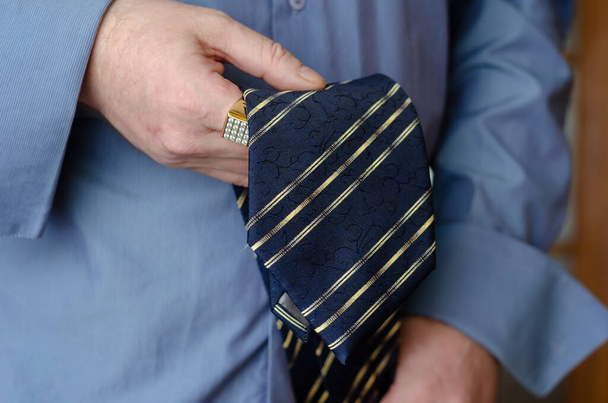 Un primer plano de una mano sosteniendo una corbata de rayas azules y amarillas. Un hombre de mediana edad con un anillo de oro en su dedo índice. Estilo de vida. De interior. - Foto, imagen