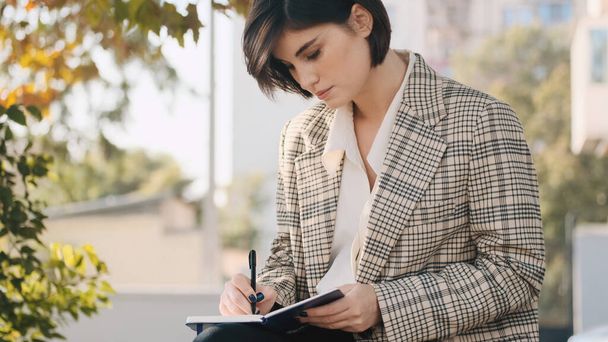 Young businesswoman sitting on street and making notes in her notebook. Attractive entrepreneur drawing up business plan for a month outdoor - Zdjęcie, obraz