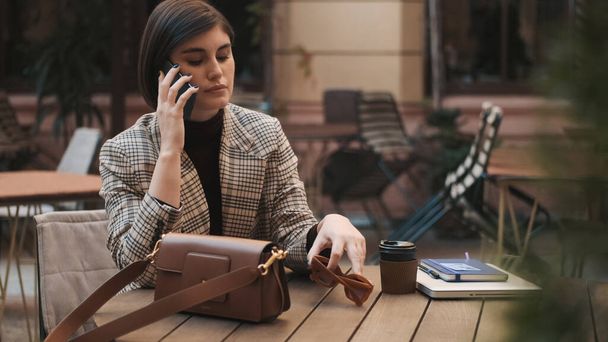 Young businesswoman sitting in cozy cafe outdoor and talking on phone looking thoughtful while solving work issues during coffee break - Фото, изображение