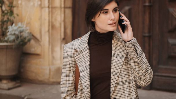 Atrakcyjna stylowa bizneswoman rozmawiająca przez telefon podczas powolnego spaceru po przytulnej ulicy - Zdjęcie, obraz