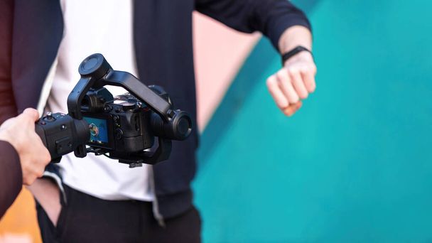 Main masculine tenant un appareil photo sur steadycam et prenant la photo d'un homme avec bracelet de fitness, fond coloré - Photo, image