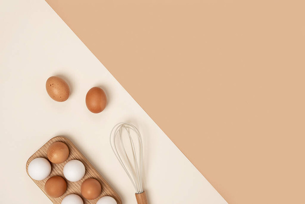 Eggs in wooden egg box and whisk on beige background. Flat lay Top view Copy space. - Фото, зображення