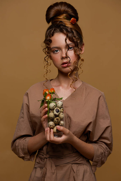 Portrait of a teenage girl. In the hair of the rose. She is holding a glass with sawn eggs and a bouquet of flowers in her hands. Unusual hairstyle. - Photo, image