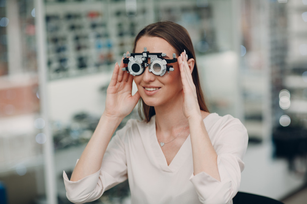 Πορτρέτο της χαρούμενης νεαρής γυναίκας κατά τη διάρκεια της εξέτασης ματιών με γυαλιά δοκιμής σε οπτικό οφθαλμίατρο - Φωτογραφία, εικόνα