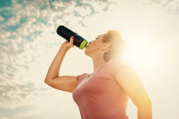 Pretty adult woman in sportswear, drinking from a shaker. Blue sky and clouds in the background. Bottom view. Sunlight. Concept of fitness and sports nutrition. - Foto, afbeelding