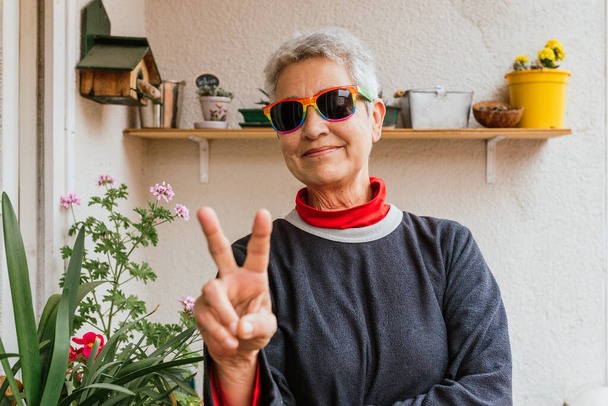 Leszbikus idős nő rövid ősz hajjal szivárvány büszkeség zászlós napszemüvegben, aki a győzelmi táblát adja elő az otthona teraszán. Emberi jogi aktivista. lgbt közösség. - Fotó, kép