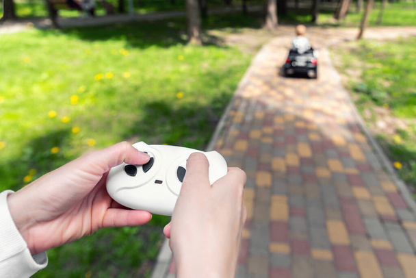 Parent hand holding remote controller of electric powered battery car with little kid boy enjoy having fun riding toy asphalt path road city park at summer day. Mother child safety and care concept - Photo, Image