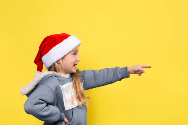 Ένα όμορφο κοριτσάκι με κόκκινο καπέλο, χαμογελάει και δείχνει το δάχτυλό της. Banner με χώρο αντιγραφής. Νέο έτος ή Χριστούγεννα έννοια - Φωτογραφία, εικόνα