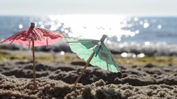 Paraguas de cóctel de papel en arena en la orilla del mar en el soleado primer día de verano - Metraje, vídeo