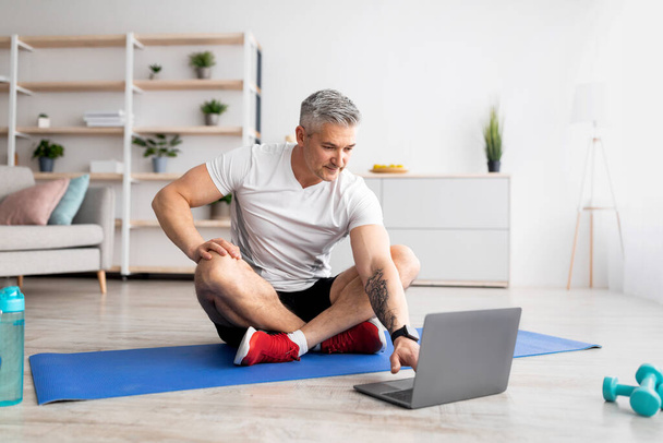 ラップトップコンピュータでオンラインスポーツのチュートリアルを見ているシニア男性,リビングルームのインテリアで自宅で運動 - 写真・画像