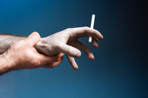 Pale crooked male hand, close-up, holding a cigarette. Healthy hand trying to quit smoking. Dark blue background. The concept of World No Tobacco Day. - Photo, Image