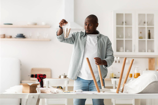 Puzzled african american man holding package with details, assembling chair with instructions in kitchen - Valokuva, kuva