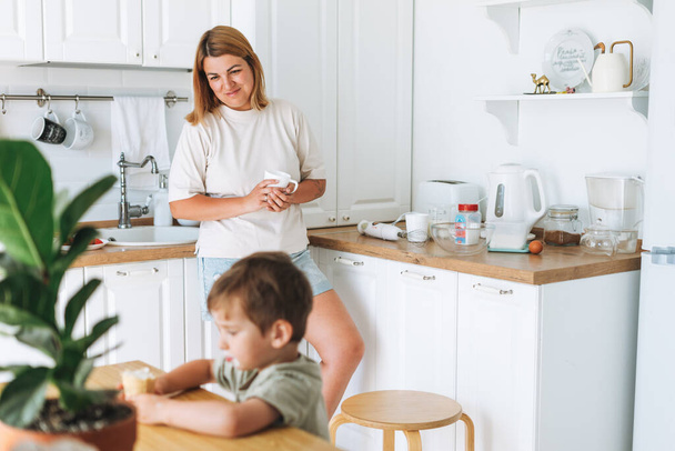 Νεαρή γυναίκα μητέρα αναζητούν ως παιδί γιο της, λαμβάνοντας πρωινό στο τραπέζι σε φωτεινή κουζίνα στο σπίτι - Φωτογραφία, εικόνα