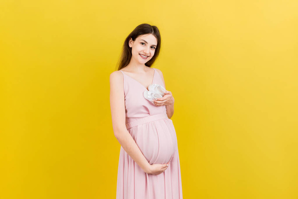 Mujer embarazada feliz sosteniendo botines de niña bebé contra su vientre en fondo de color. Feliz embarazo. Copiar espacio. - Foto, imagen