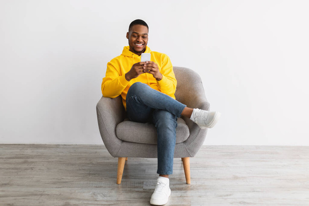 African Guy Using Smartphone Sitting In Chair Over Gray Wall - Photo, Image